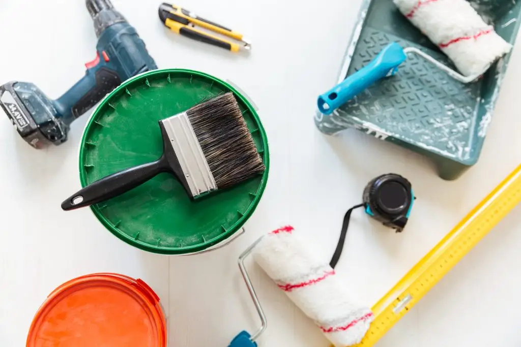 Verschiedene Renovierungswerkzeuge wie Pinsel, Farbroller, Bohrmaschine, Cutter, Maßband und Farbeimer auf hellem Untergrund